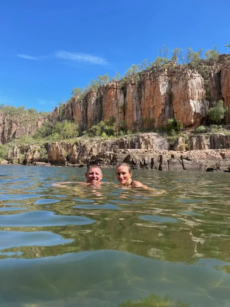 Two people swimming in a river with a rocky cliff and green trees in the background, under a clear blue sky.