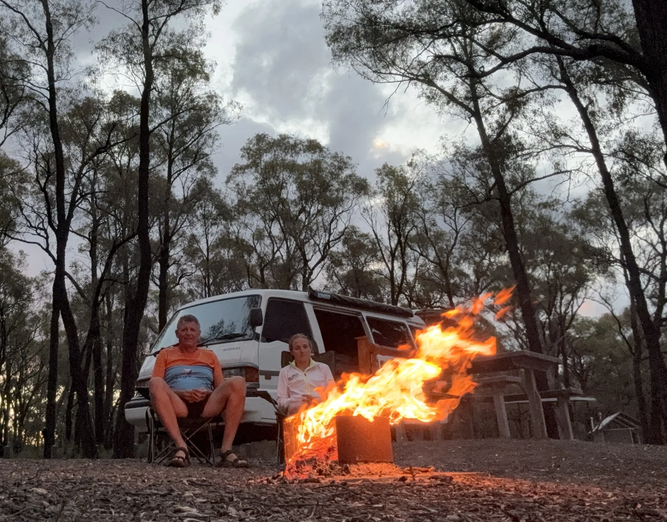 A man and a woman sitting in camping chairs near a campfire in a wooded area during dusk, with a van in the background and trees surrounding them.