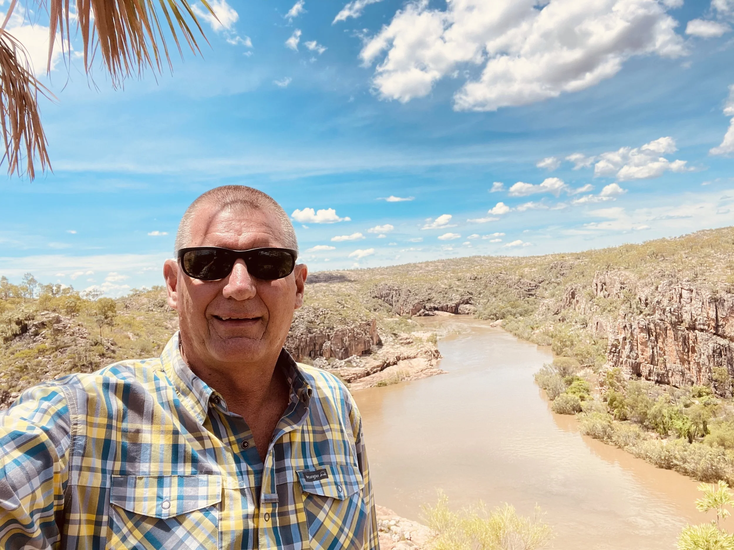 A man with short gray hair wearing sunglasses and a yellow plaid shirt takes a selfie by a river in a canyon with rocky cliffs on both sides, under a blue sky with scattered clouds.