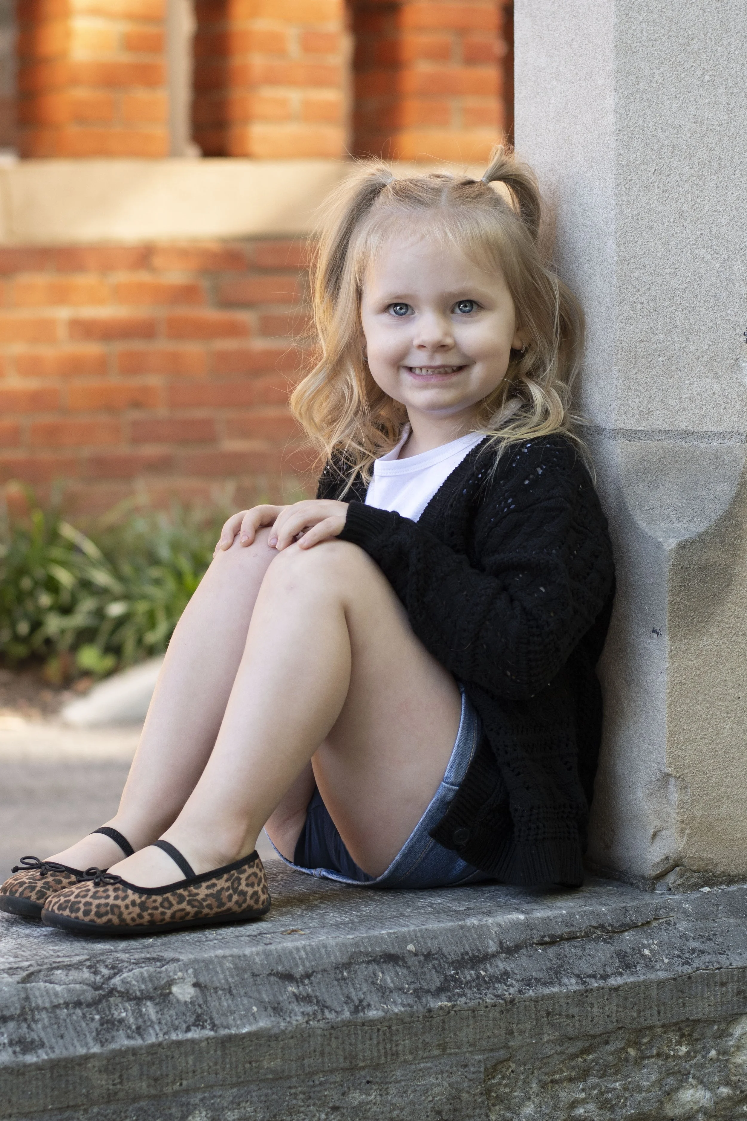 A young girl with blonde hair, blue eyes, and a cheerful smile, sitting on a concrete ledge outside a building with red brick walls and a gray corner.