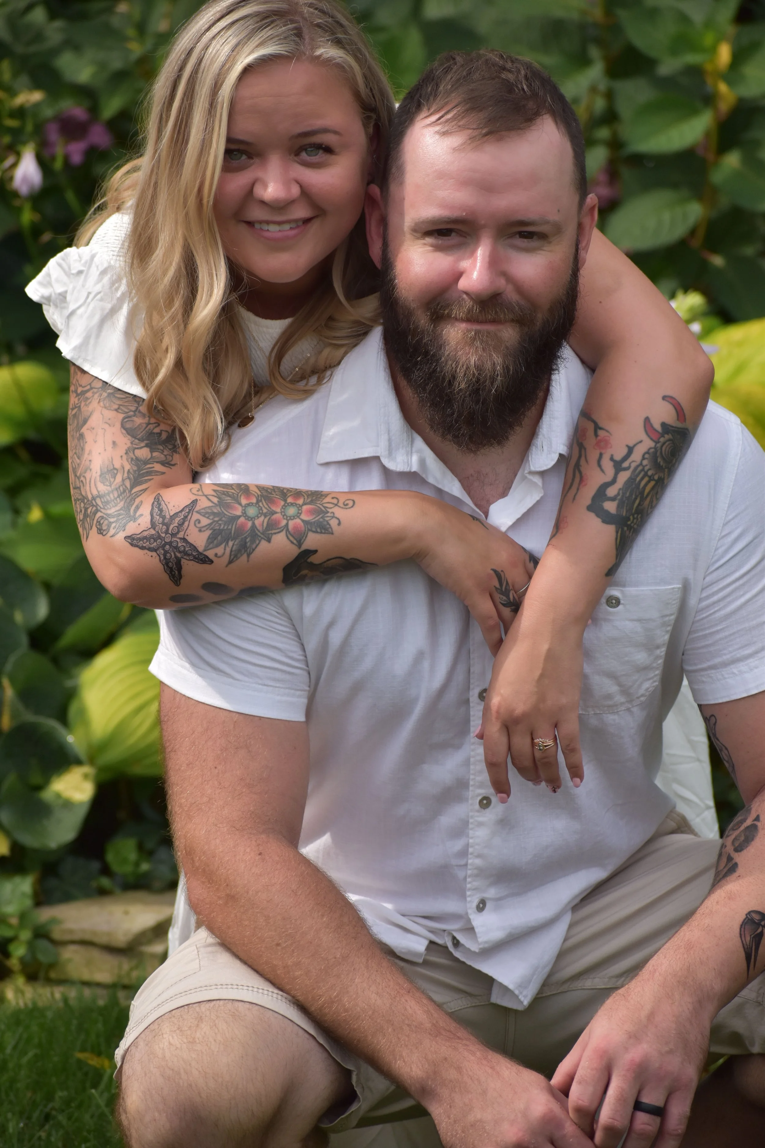 A smiling woman with blonde hair and tattoos on her arms is giving a piggyback ride on a man with a beard, sitting outdoors with green foliage in the background.
