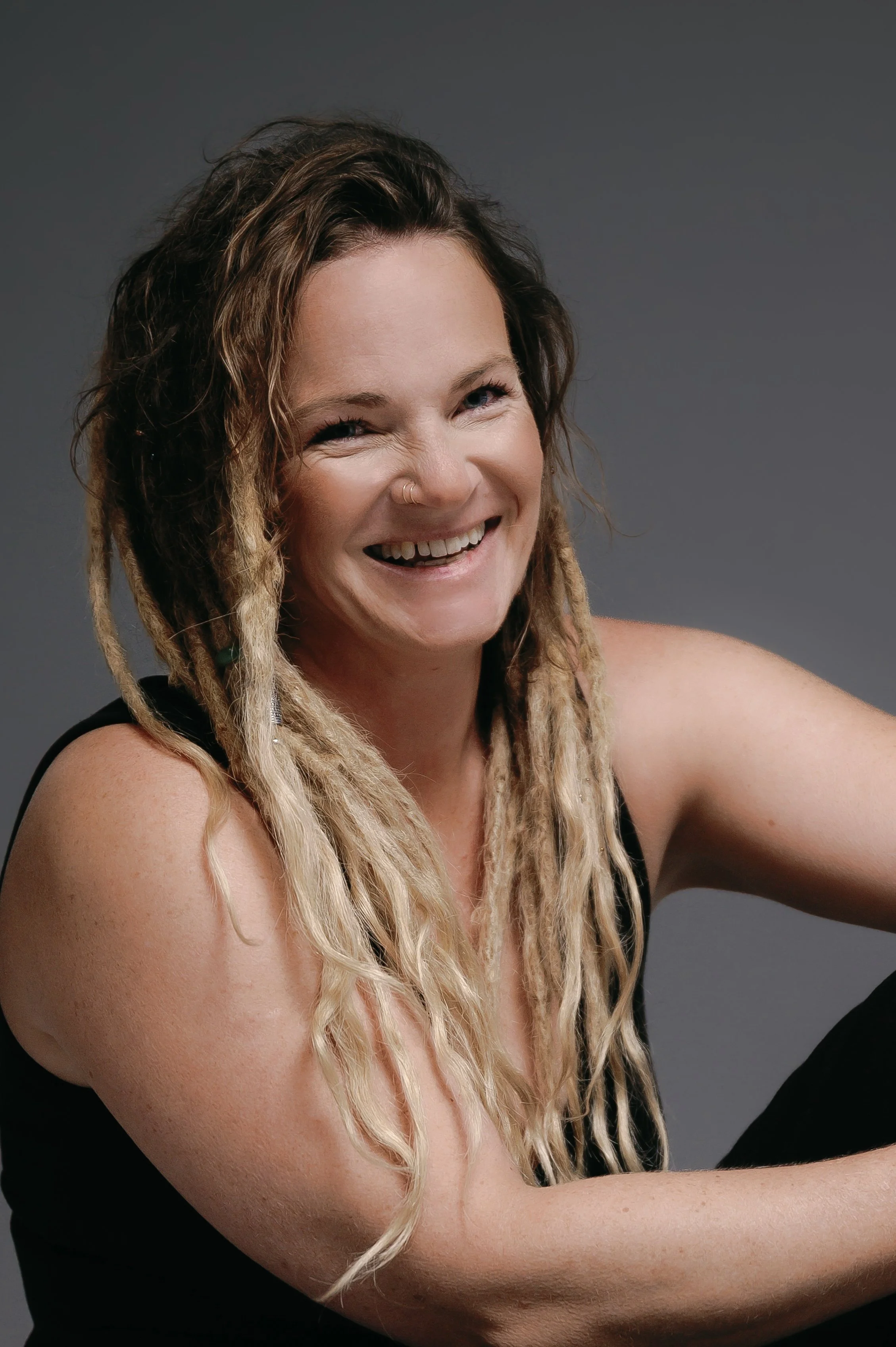 Close-up of a woman with dreadlocks smiling and lifting her arm, set against a grey background.