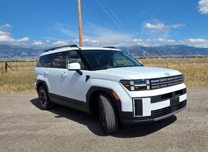 A white SUV parked on a gravel road in open countryside with mountains in the background and a utility pole nearby.