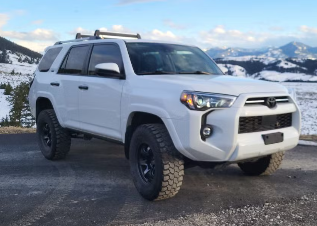 White Toyota 4Runner SUV parked on a mountain road with snow-covered mountains in the background.
