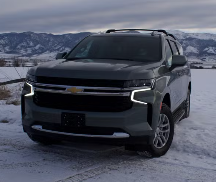 A dark gray Chevrolet SUV parked on a snowy landscape with mountains in the background and overcast sky.