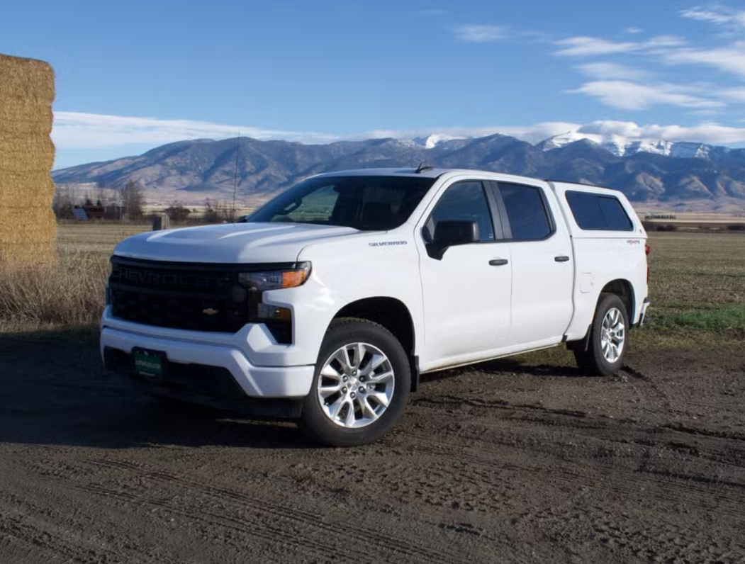 A white Chevrolet pickup truck parked on a dirt road with mountains in the background and a stack of hay bale on the left.
