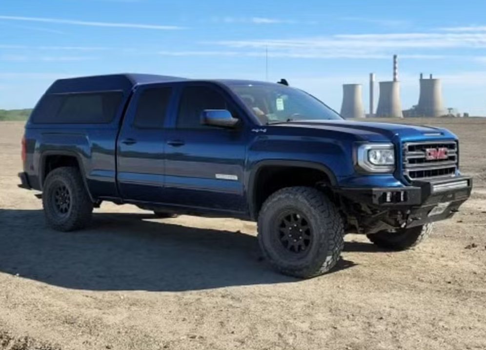 A dark blue GMC pickup truck with a camper shell parked on a dirt area near a nuclear power plant with cooling towers in the background under a blue sky.