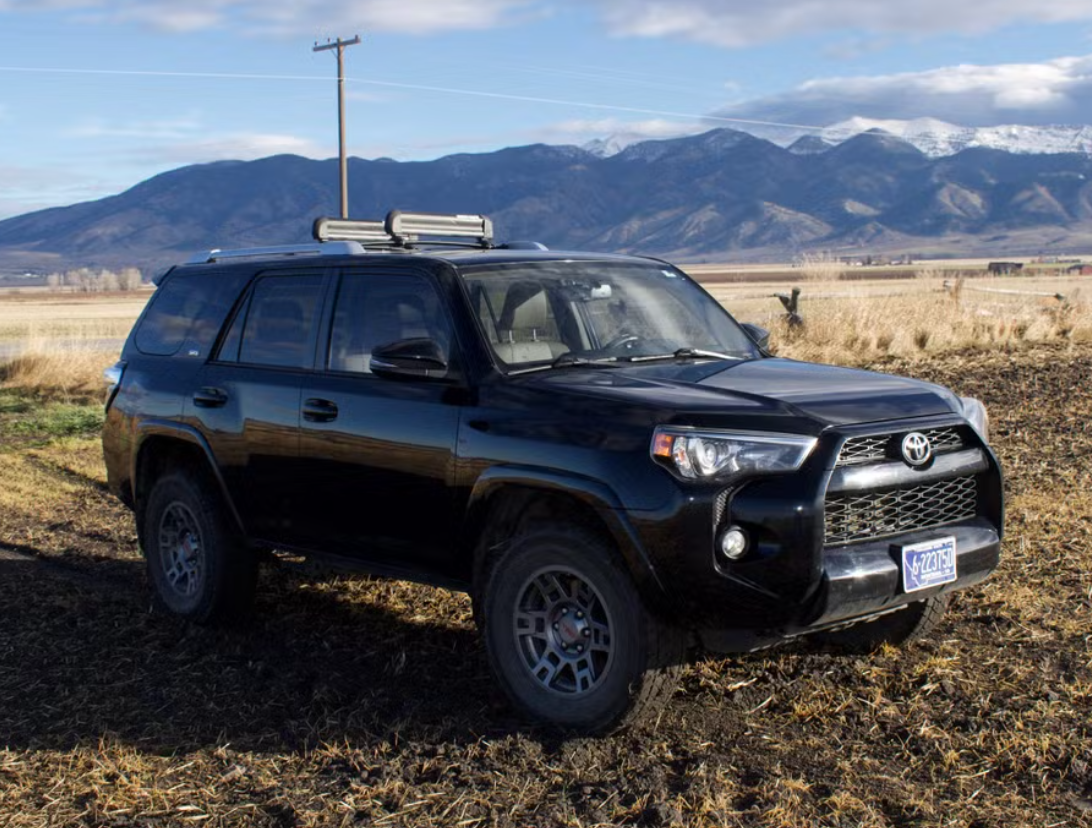 Black Toyota 4Runner SUV parked in a rural field with mountains in the background, a utility pole nearby, and a partly cloudy sky.