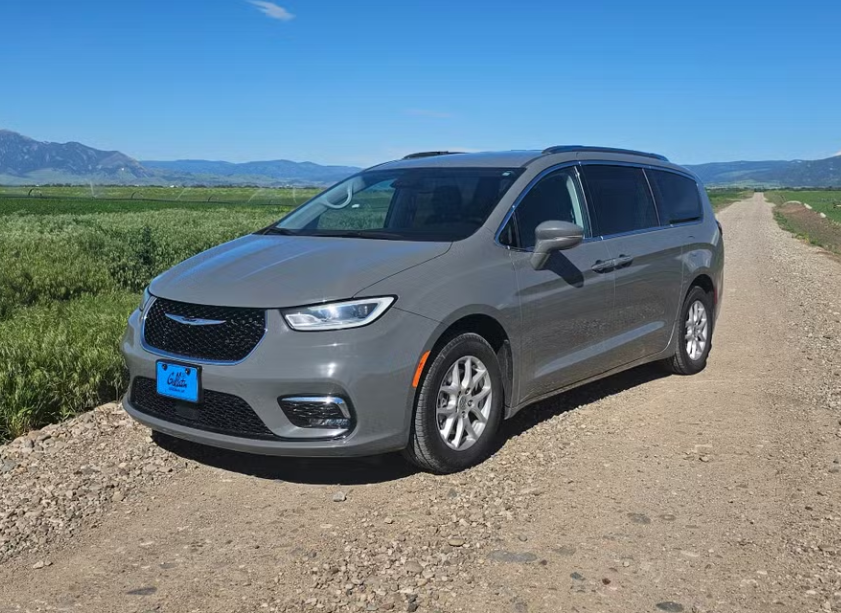 Gray minivan parked on a dirt road in a rural area with green fields and mountains in the background under a clear blue sky.