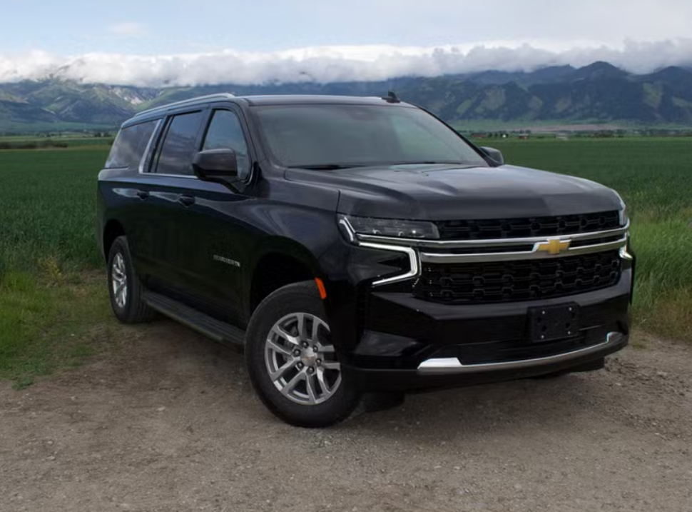 Black Chevrolet SUV parked on dirt road in a green field with mountains and cloudy sky in the background.