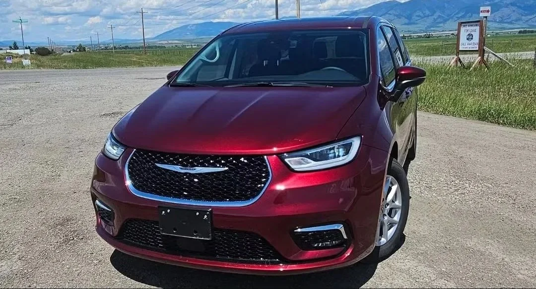 Red Chrysler minivan parked on gravel with open fields and mountains in the background.