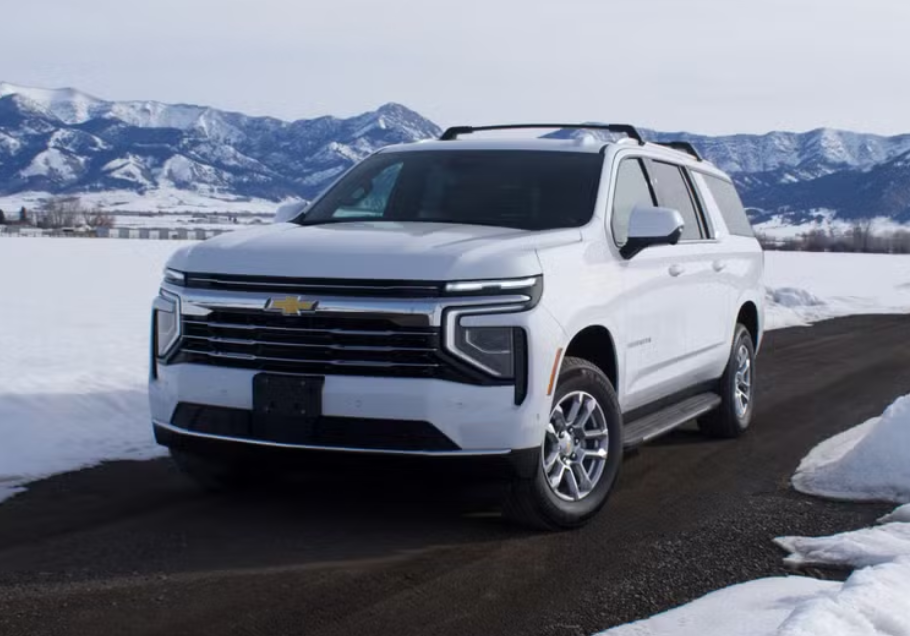 A white Chevrolet SUV parked on a dark paved road in a snowy landscape with snow-capped mountains in the background.