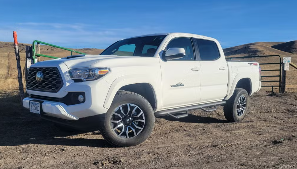 A white Toyota pickup truck parked on a dirt area with hills and a metal gate in the background.
