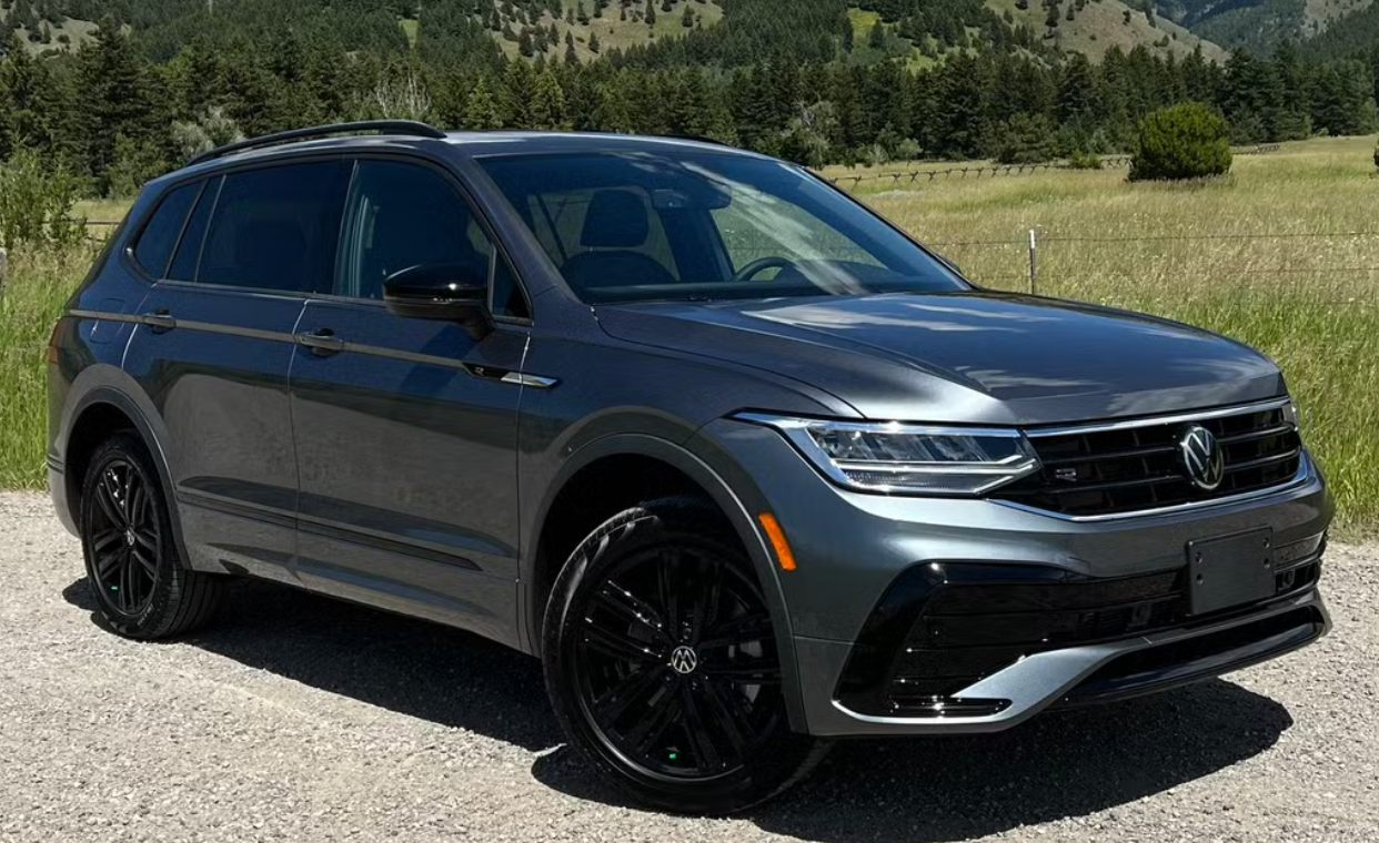 A gray Volkswagen SUV parked outdoors on a gravel path, with green grass, trees, and mountains in the background.