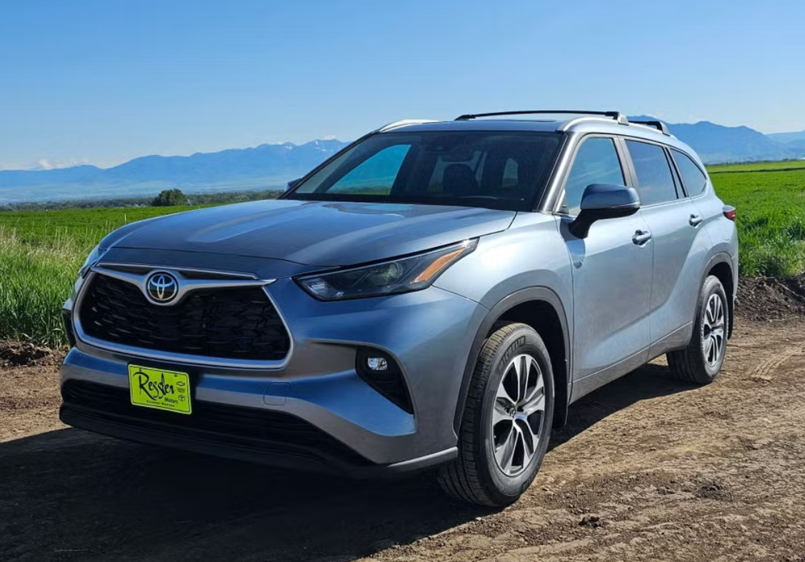 A silver Toyota RAV4 parked on a dirt road with a green field and mountains in the background under a blue sky.