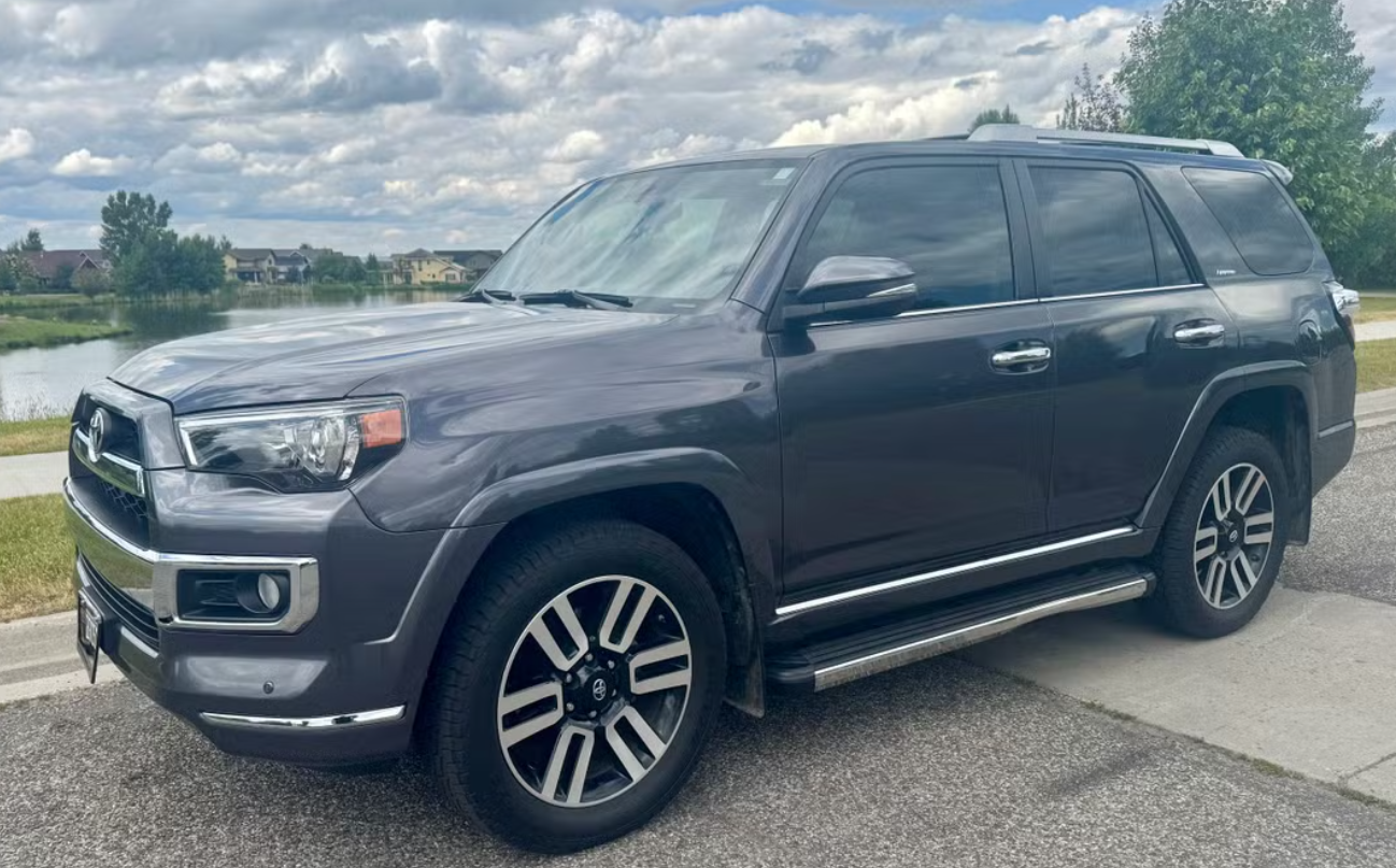 A black Toyota 4Runner SUV parked at the side of a road near a body of water with houses and trees in the background and a cloudy sky.