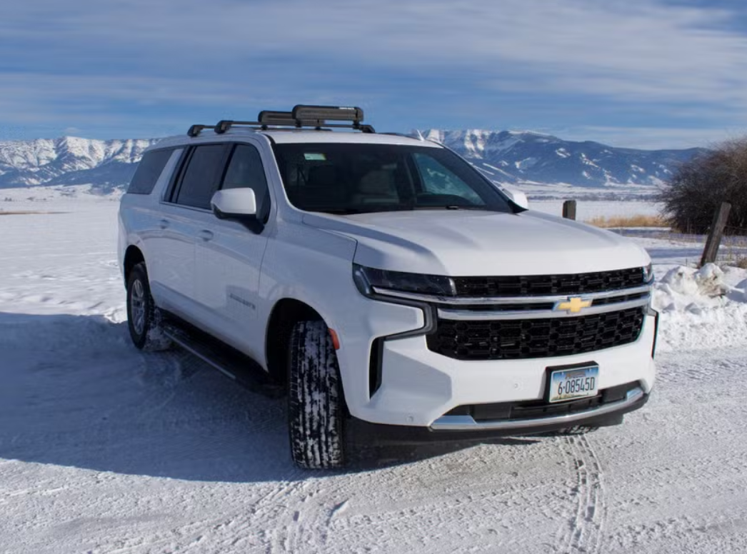 White Chevrolet SUV parked on snow-covered ground with mountains in the background and a partly cloudy sky.