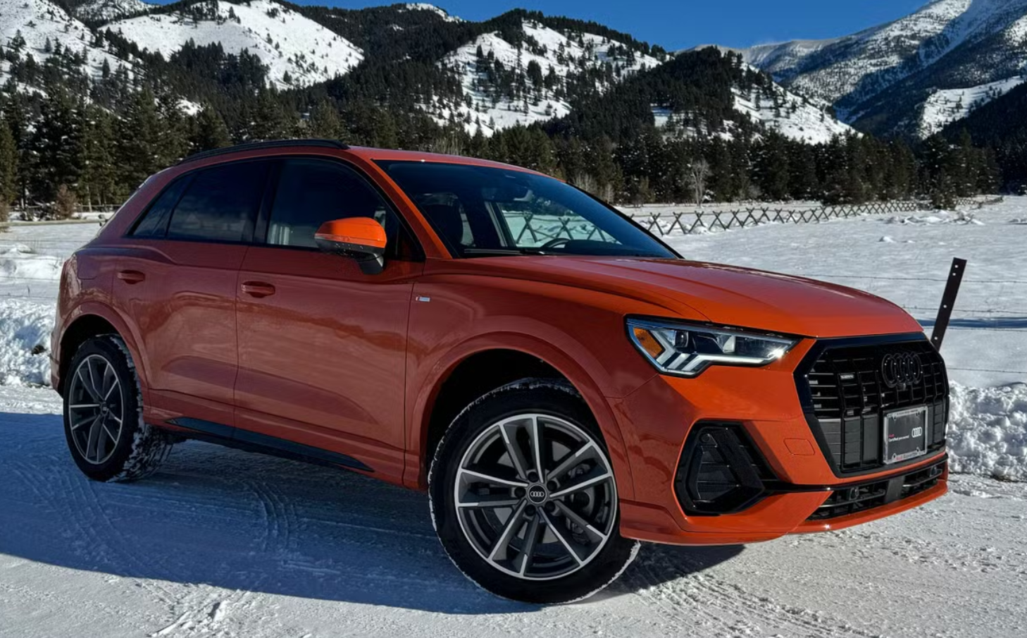 An orange Audi SUV parked on a snowy landscape with snow-covered mountains and trees in the background.