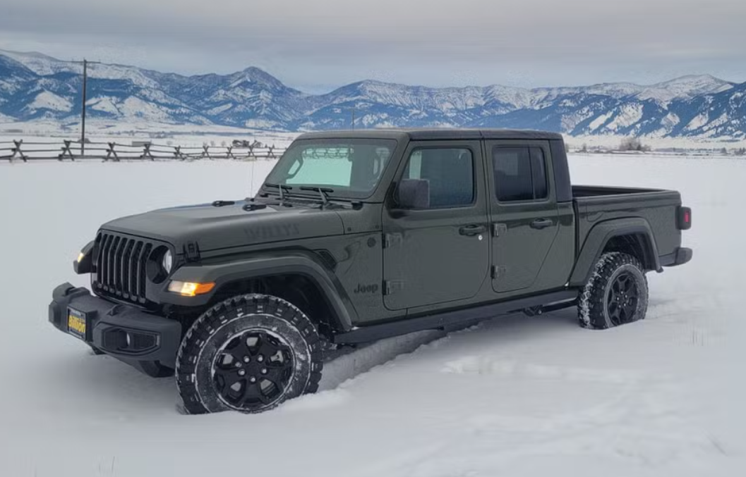 A dark green Jeep Gladiator pickup truck parked in the snow with mountain ranges in the background.
