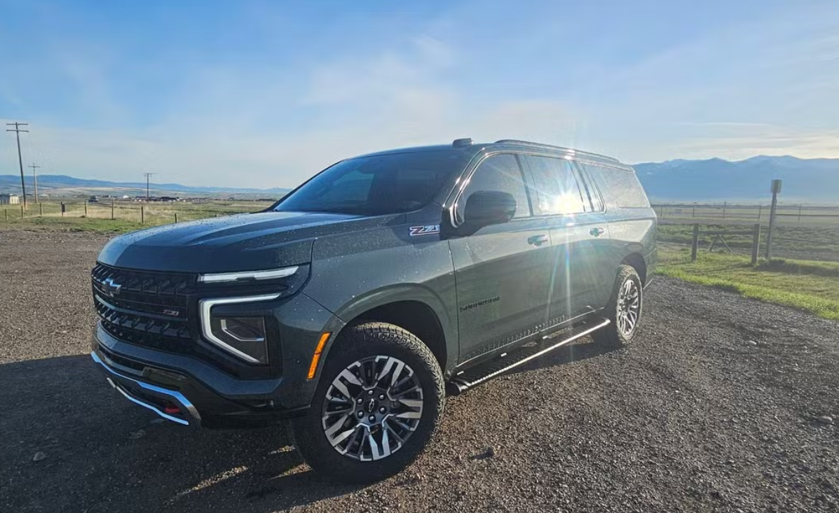 A dark gray Chevrolet SUV parked on a dirt lot with open fields and mountains in the background, sunlight reflecting off the vehicle.
