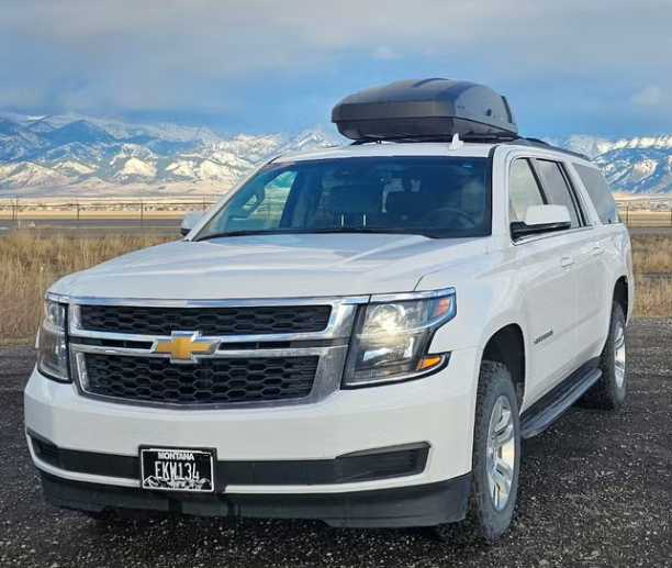 White Chevrolet SUV with a cargo box on top, parked on a gravel surface in front of snowy mountains under a partly cloudy sky.