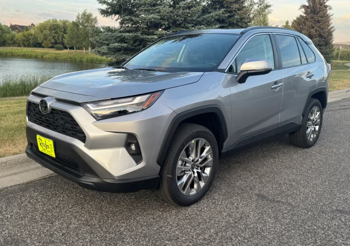 A silver Toyota RAV4 parked on a paved street with a grassy area and trees in the background during daylight.