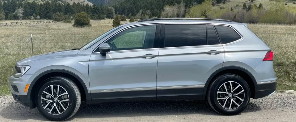 Silver SUV parked on a gravel road near a grassy field and trees with hills in the background.