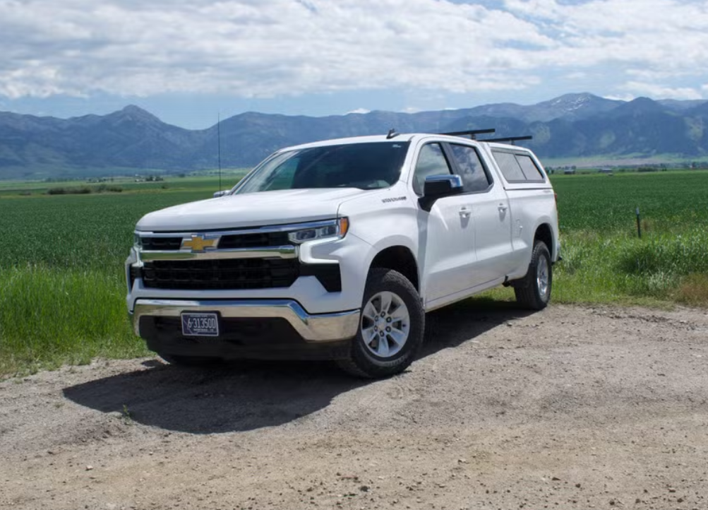 White Chevrolet pickup truck parked on a dirt road with green fields and mountains in the background.