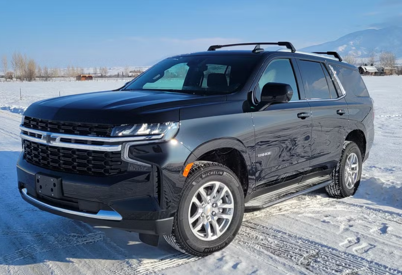 Black and gray Chevrolet Tahoe SUV parked in a snowy landscape with mountains and clear blue sky in the background.