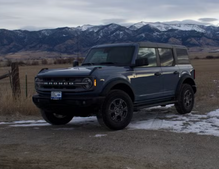 A dark gray Ford Bronco SUV parked on a dirt road with snow patches, with a rural landscape and mountain range with snow-capped peaks in the background.