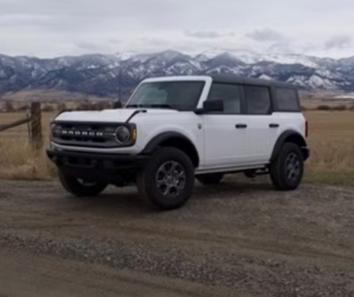A white off-road vehicle, likely a Ford Bronco, parked on a dirt road with mountains in the background.