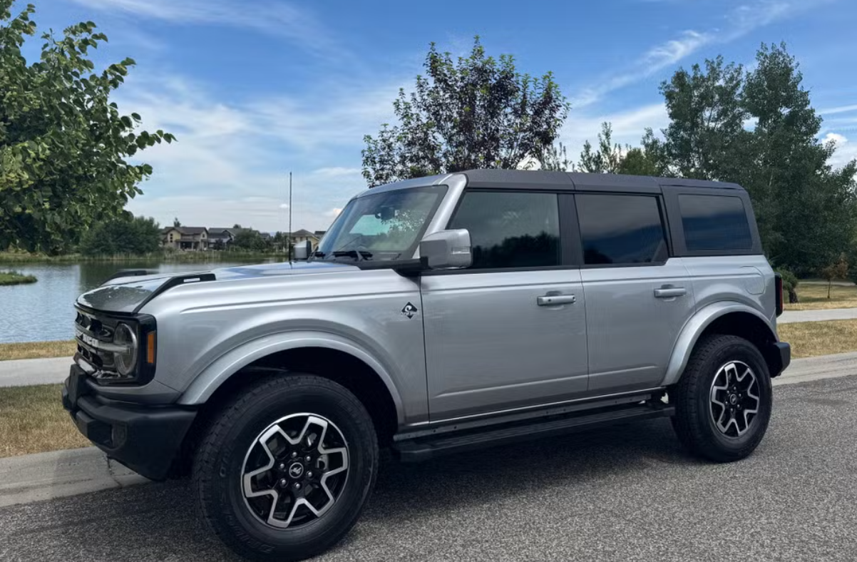 A silver SUV parked on the side of a road near a body of water with trees and houses in the background on a clear day.