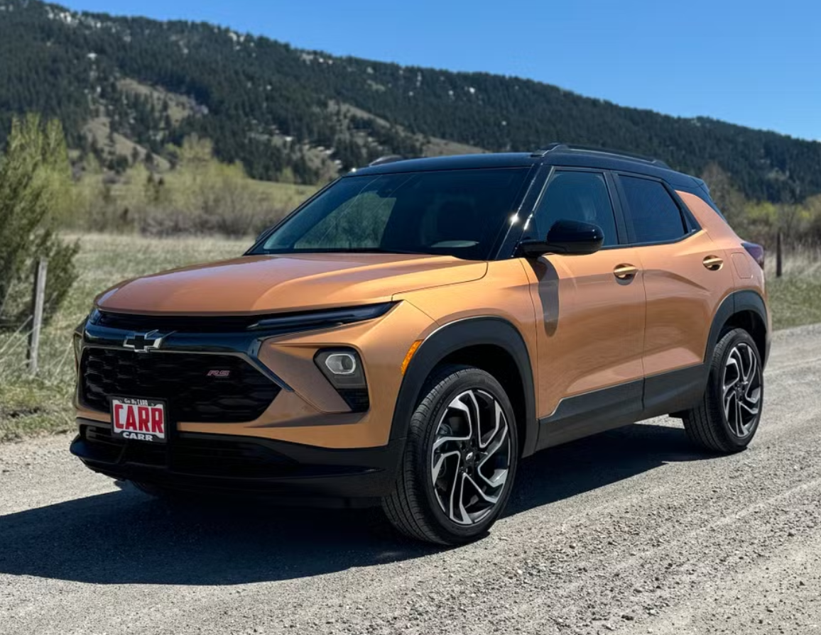 Orange Chevrolet SUV parked on a gravel road with a mountainous landscape and blue sky in the background.