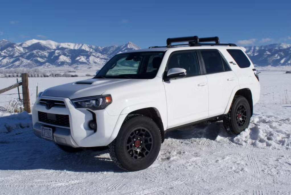 White SUV parked on snow-covered ground with snowy mountains in the background.