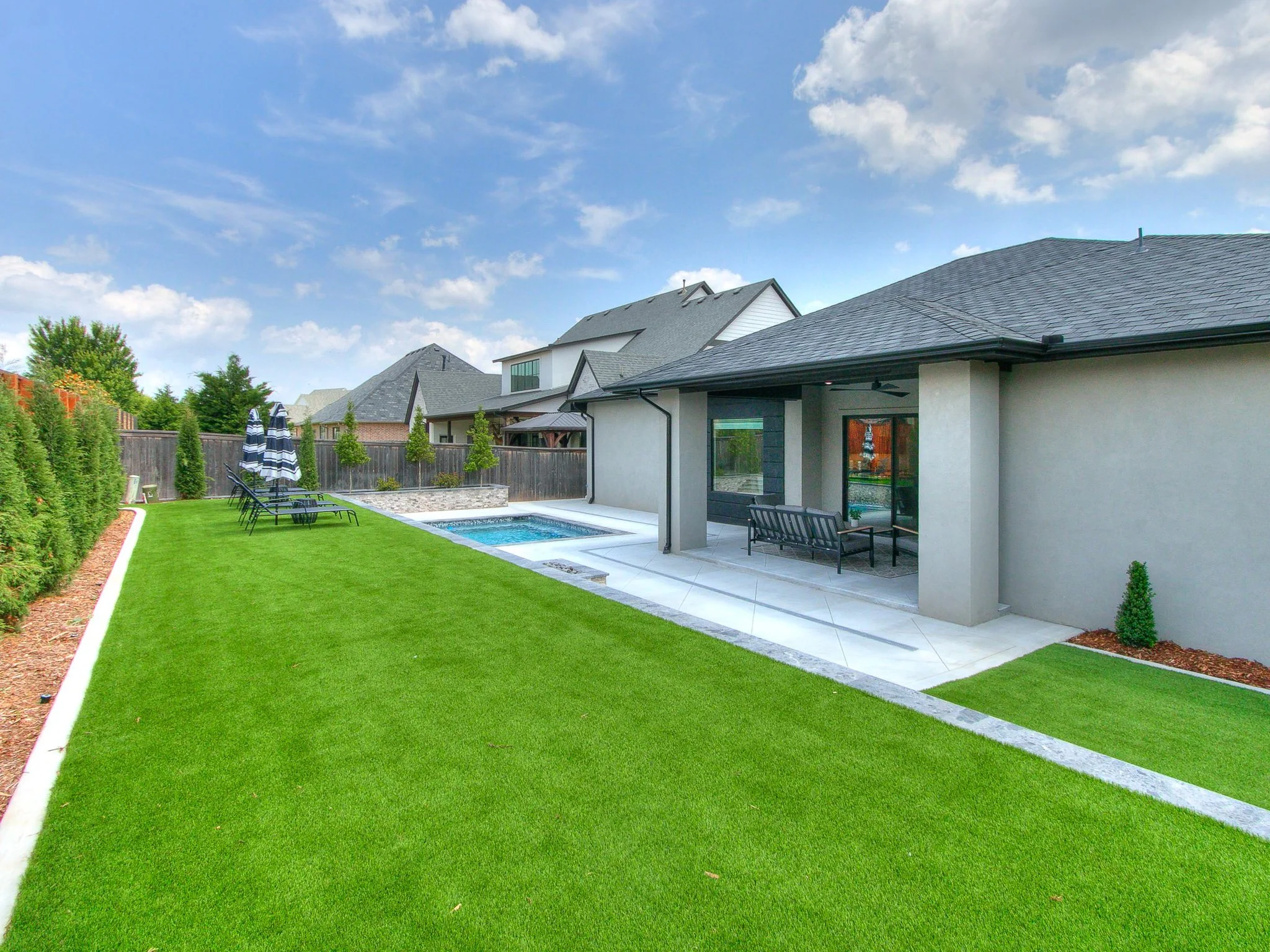 Backyard with a green lawn, patio, small pool, outdoor seating, and a fence, under a partly cloudy sky.