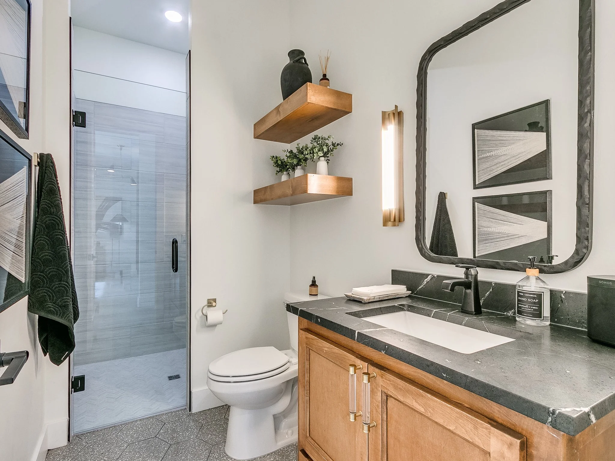 Modern bathroom with black and white decor, wooden vanity, black countertop, framed mirror, glass shower door, toilet, and wall shelves with decorative items.