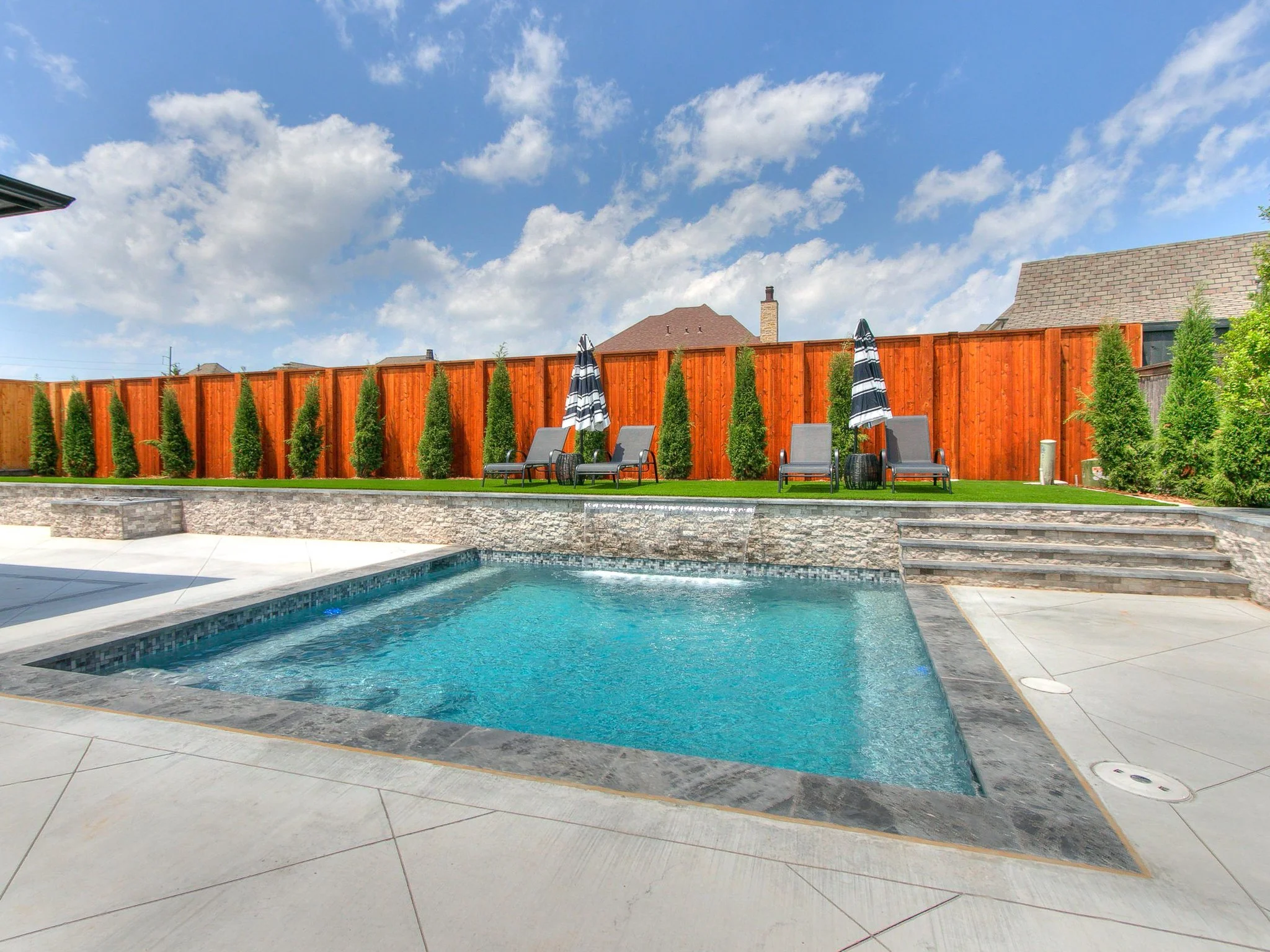 Backyard swimming pool with stone border, surrounded by a concrete deck, with a raised lawn area with four lounge chairs and umbrellas, separated by a wooden fence with tall small trees, under a partly cloudy sky.