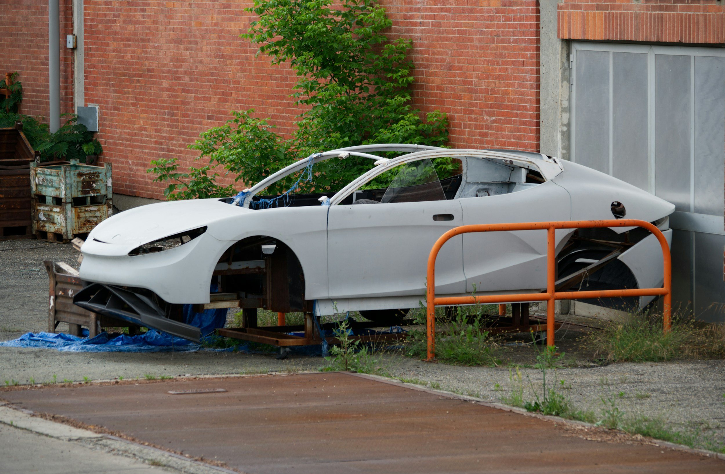 A white sports car body in the process of assembly, elevated on a platform outside a building with red brick walls and green plants.