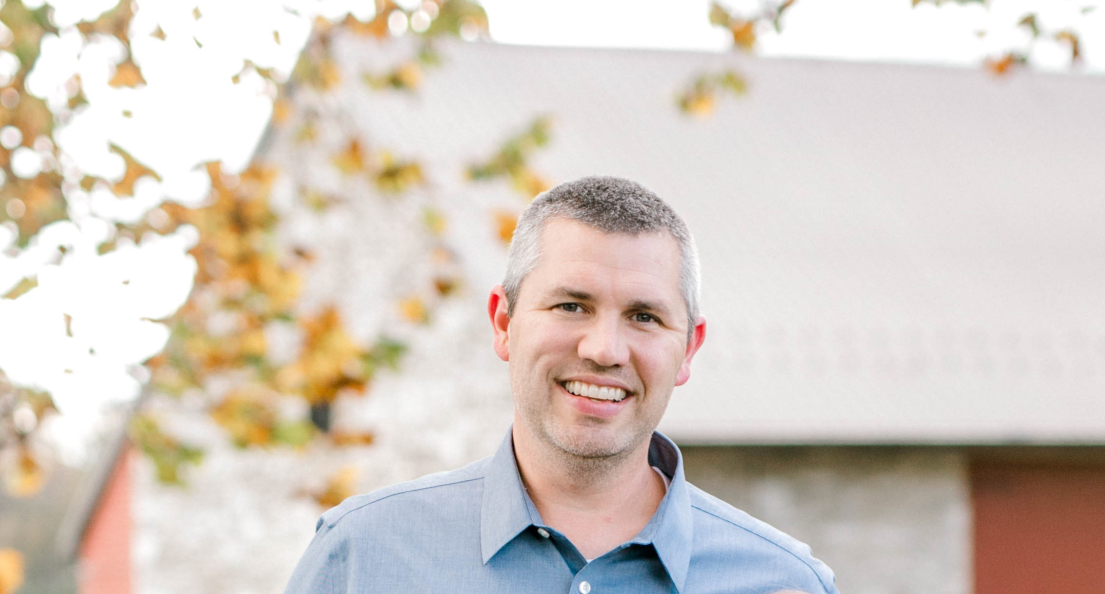 A smiling man with short gray hair wearing a blue button-up shirt standing outdoors with trees and a building in the background.
