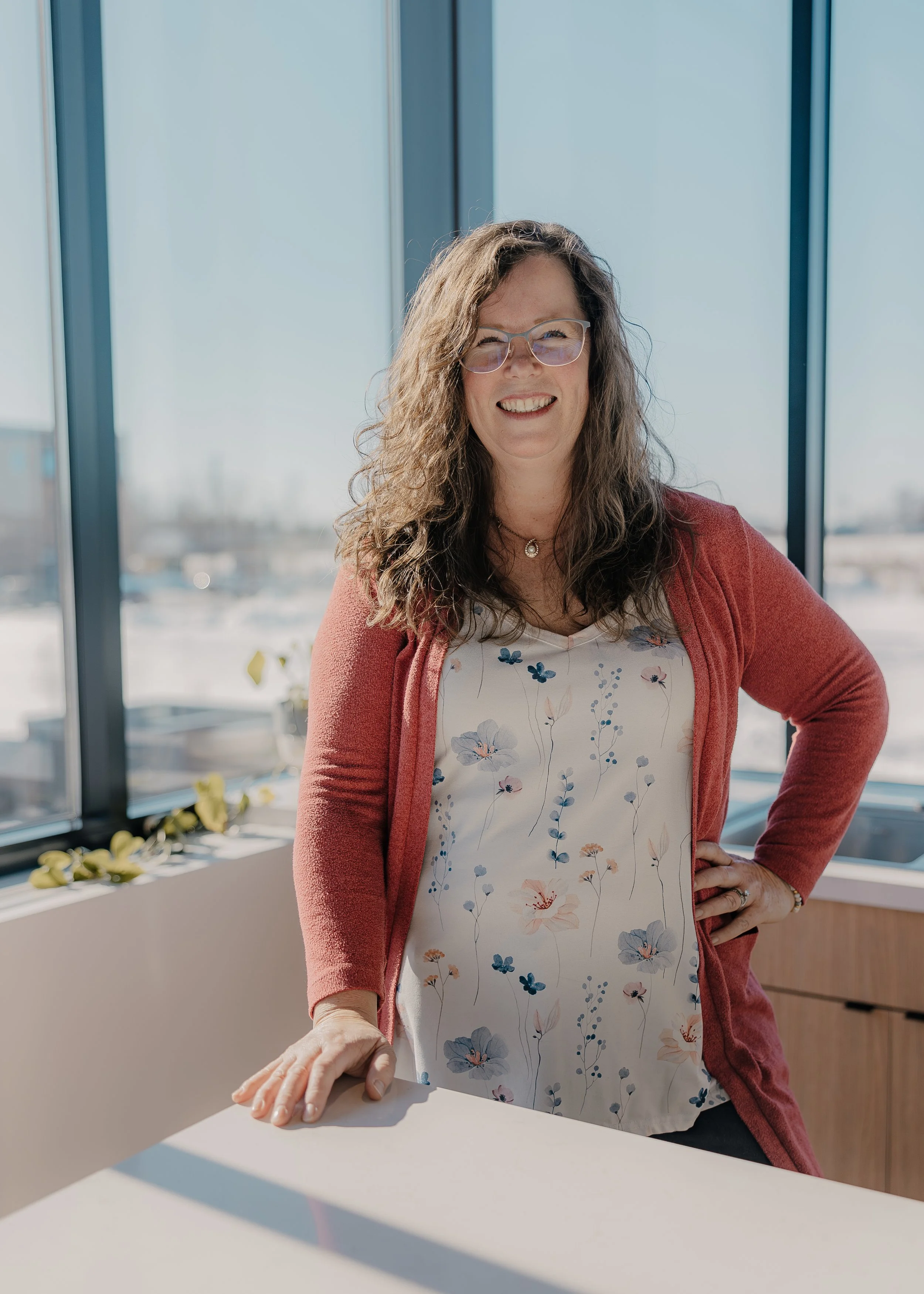 A woman with curly hair and glasses smiling, standing indoors near large windows with cityscape in the background, wearing a floral dress and a red cardigan.