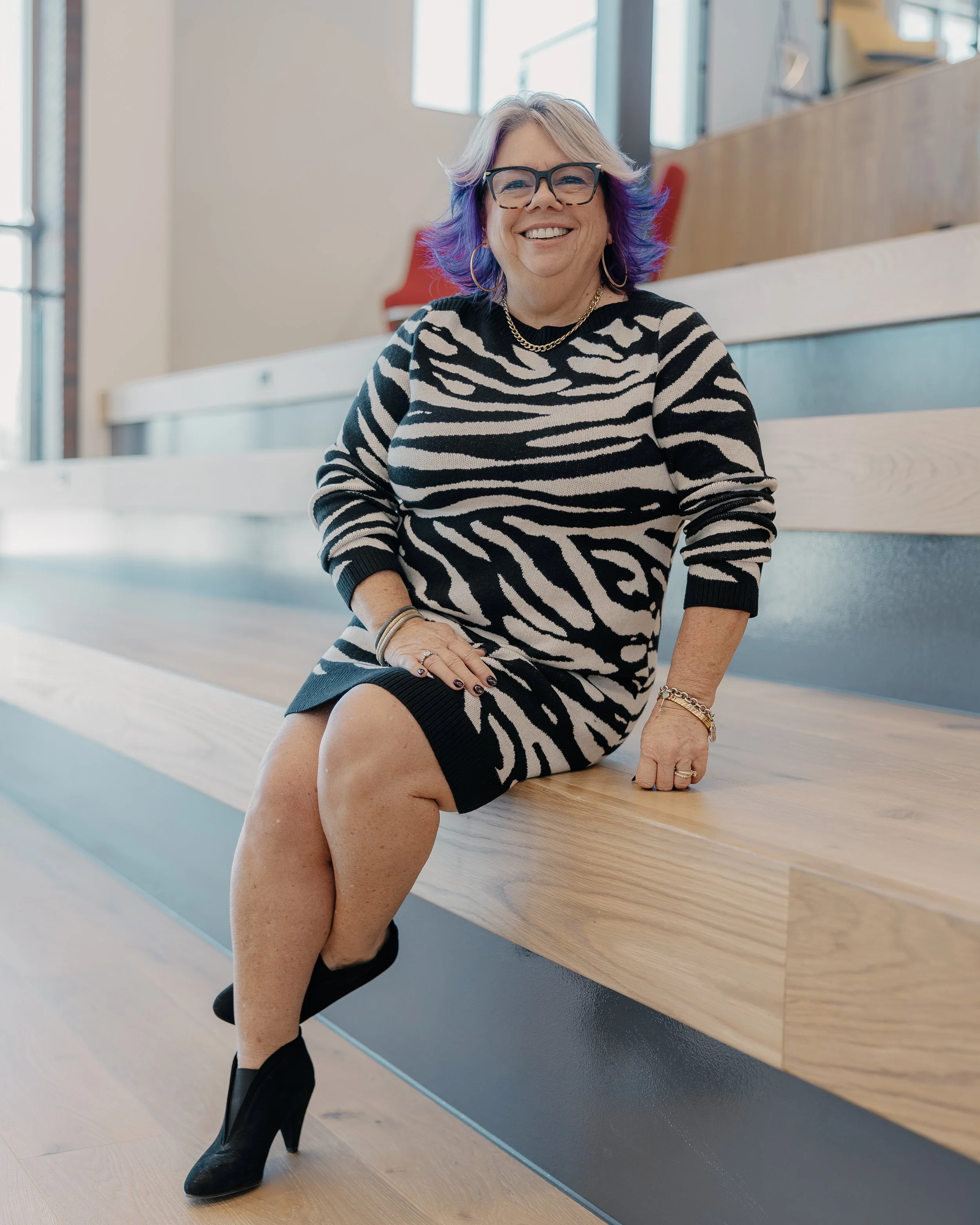 A woman sitting on a wooden bench in a modern, well-lit indoor space. She has purple and gray hair, is wearing black glasses, a black and white zebra print dress, and black high-heeled boots. She is smiling and wearing gold jewelry.