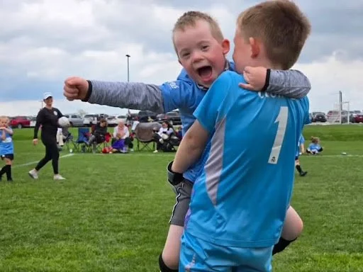 Two young boys, one in a blue sports jersey with the number 1, hugging and smiling on a grassy field during a sporting event. The boy in the jersey is lifting the other boy, who appears excited and happy. There are other people and cars in the background under a cloudy sky.