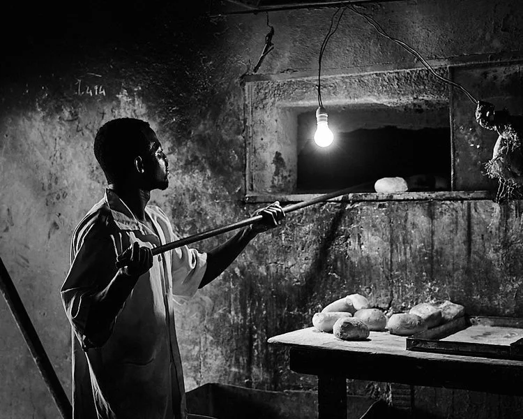 A young man in a modest kitchen, holding a stick and inspecting bread loaves on a table, illuminated by a hanging light bulb.