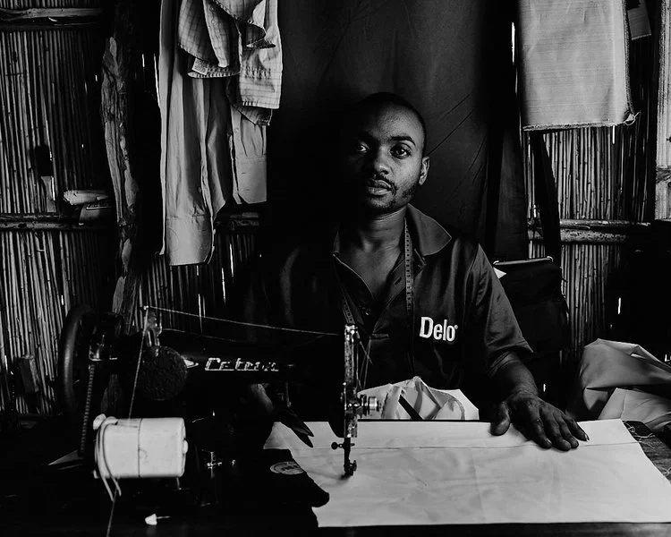 Man sitting at a worktable with sewing machine in a rustic workspace with hanging clothes and bamboo walls