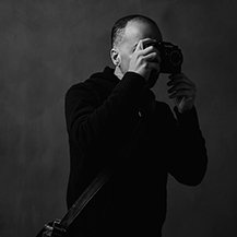 A person taking a photo with a camera, wearing a dark hoodie or jacket, in a black and white setting.