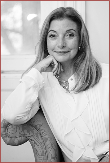 Black and white portrait of a woman with shoulder-length hair, wearing a light-colored blouse, sitting in a chair with her hand resting on her chin, smiling softly.