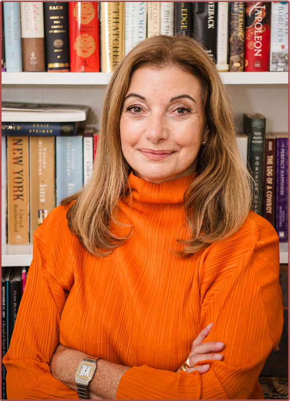 A woman with blonde hair and a warm smile, wearing an orange blouse, standing in front of a bookshelf filled with various books.