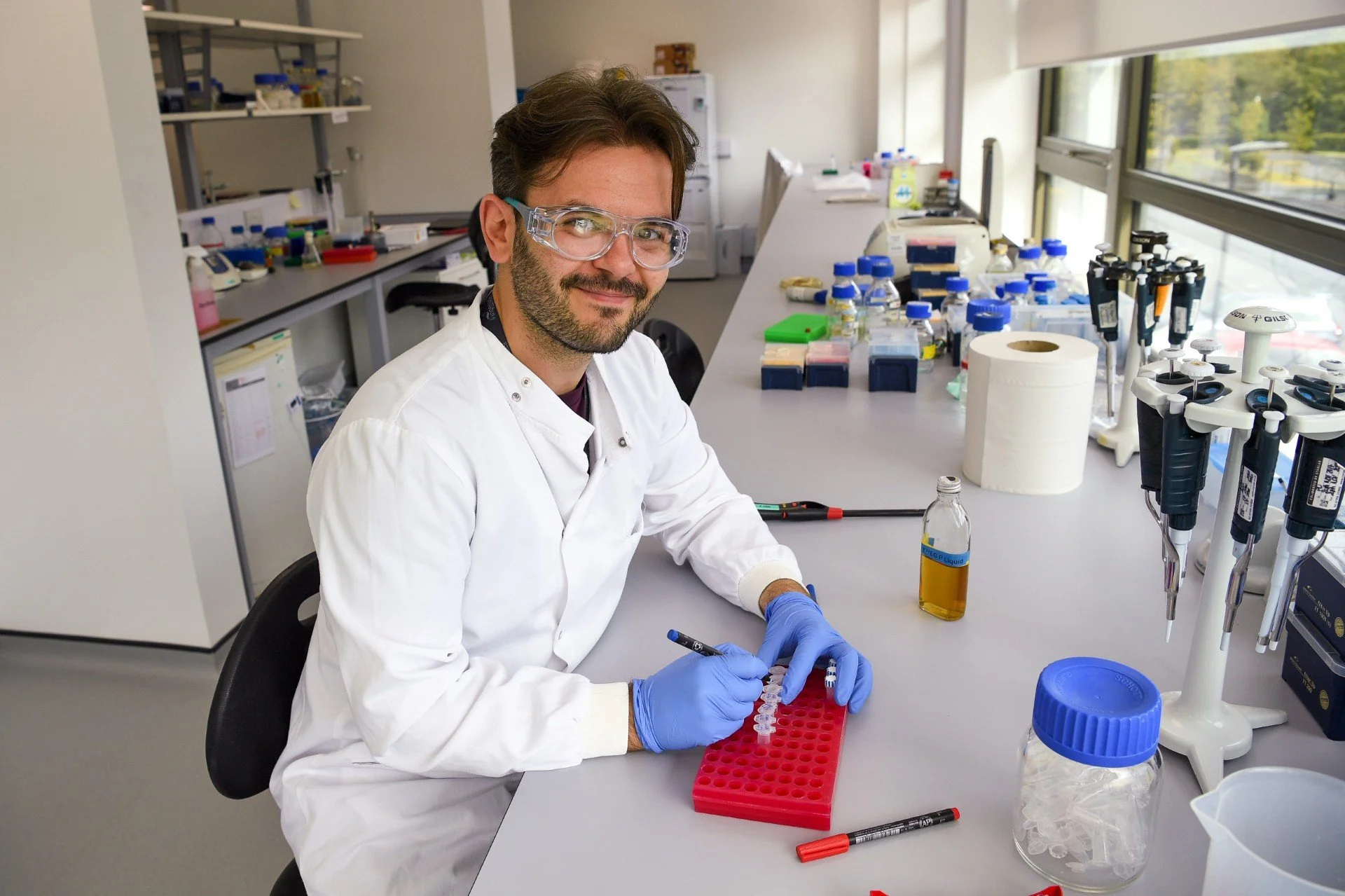 Senior Scientist in Molecular Biology  Jack Kay wearing a lab coat and safety goggles, sitting at a lab bench with a set of samples in test tubes