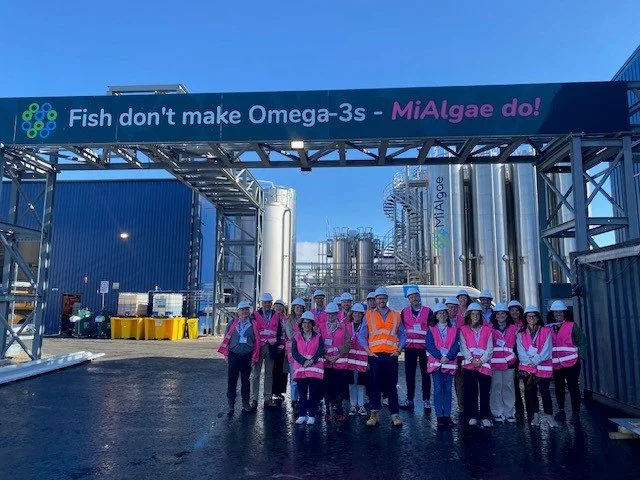 ECIV consortium delegates in pink high vis and hard hats standing outside MiAlgae's facility at Grangemouth