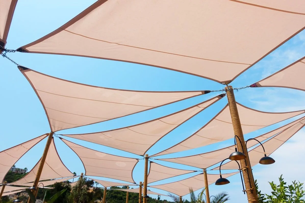 Sombrillas de tela beige colgadas en soporte de madera, con lámparas negras colgantes, bajo un cielo azul.